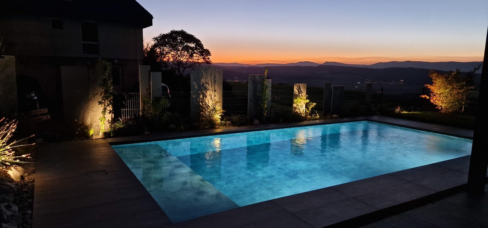 Piscine béton éclairée au crépuscule, eau turquoise lumineuse et coucher de soleil sur les collines