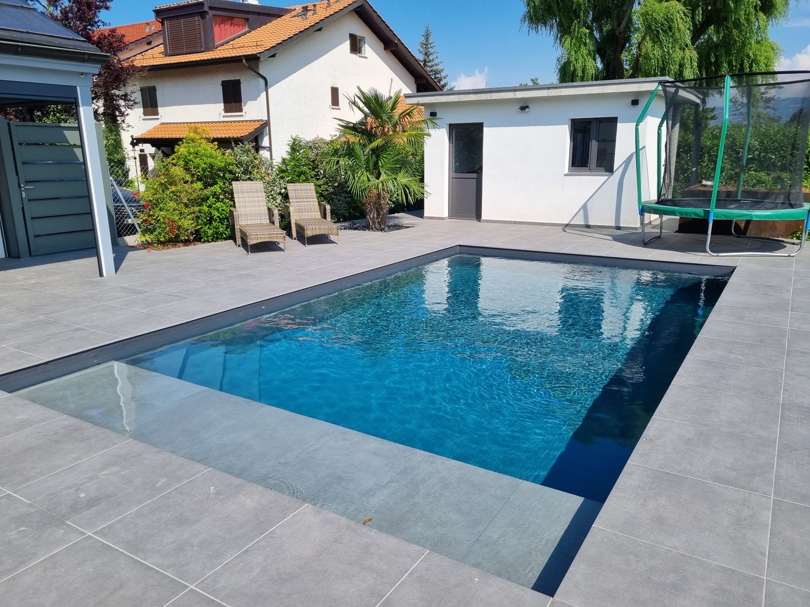 Piscine béton à l'eau bleu clair avec escalier intégré, terrasse anthracite et pool house blanc