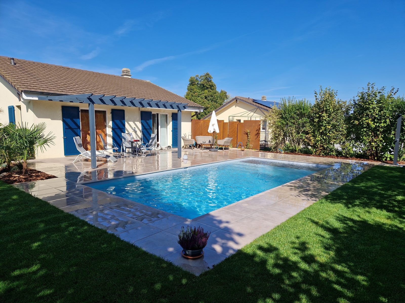 Piscine coque rectangulaire à l'eau turquoise devant une maison avec pergola, plage en carrelage beige et gazon