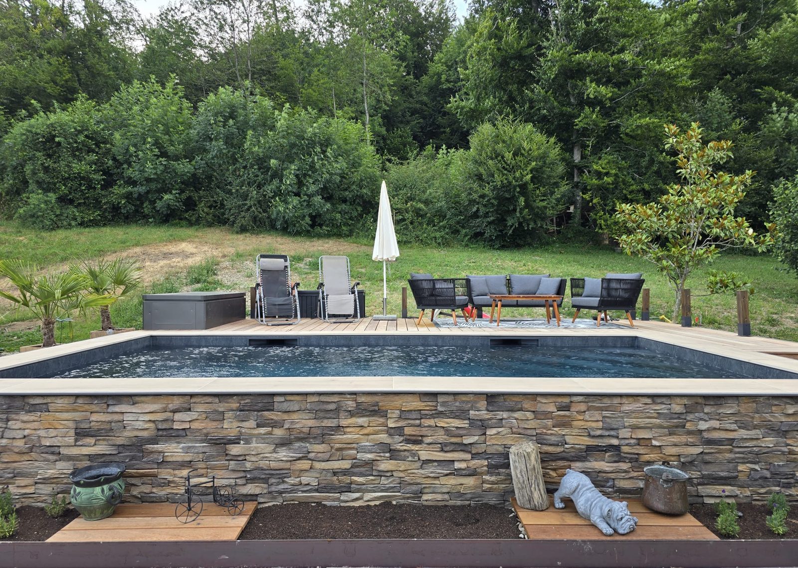 Piscine hors-sol béton habillée de parement pierre naturelle, margelles claires et salon de jardin