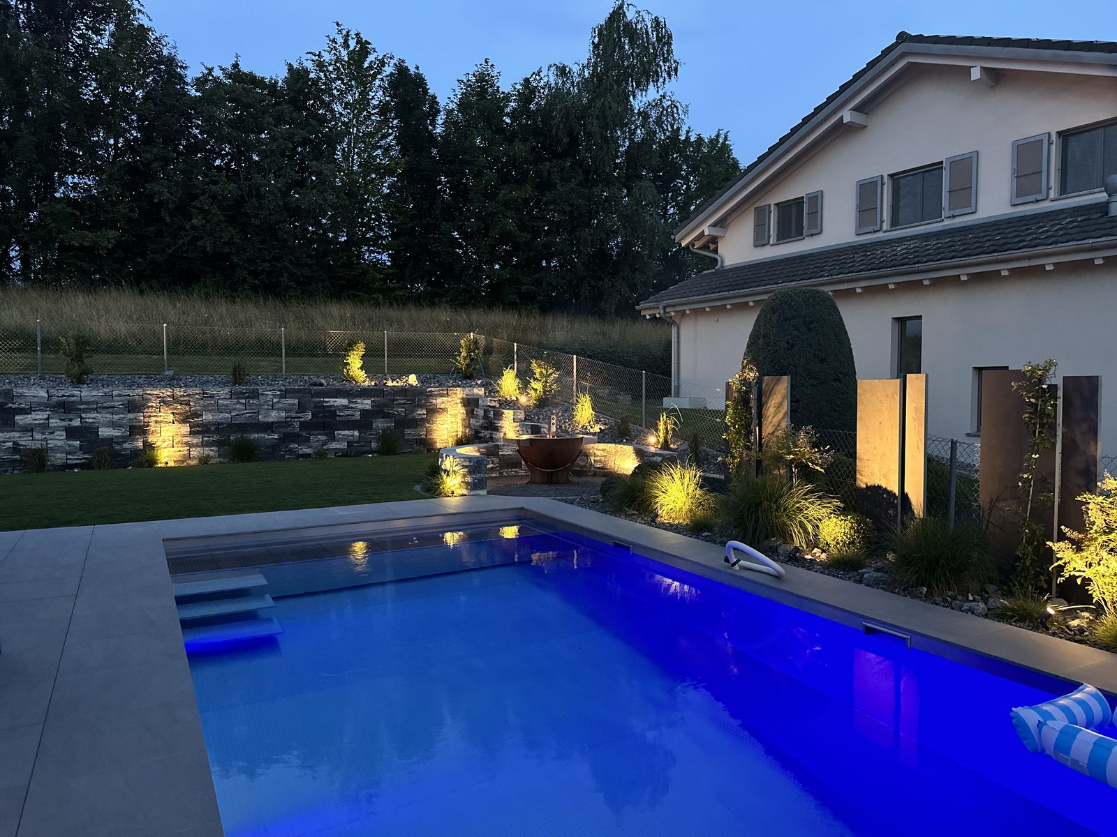 Piscine inox éclairée en bleu au crépuscule, jardin paysager illuminé et villa